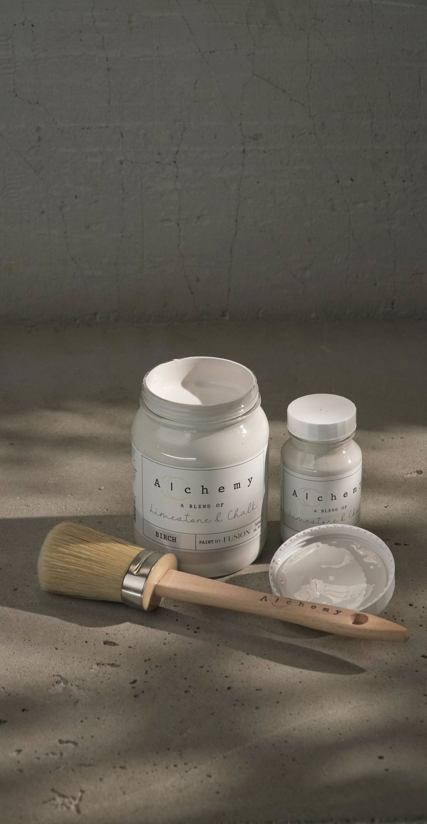Botes Alchemy by Fusion Birch 500ml y 120ml con brocha sobre fondo de cemento texturizado, pintura a la tiza off-white con subtonos naturales