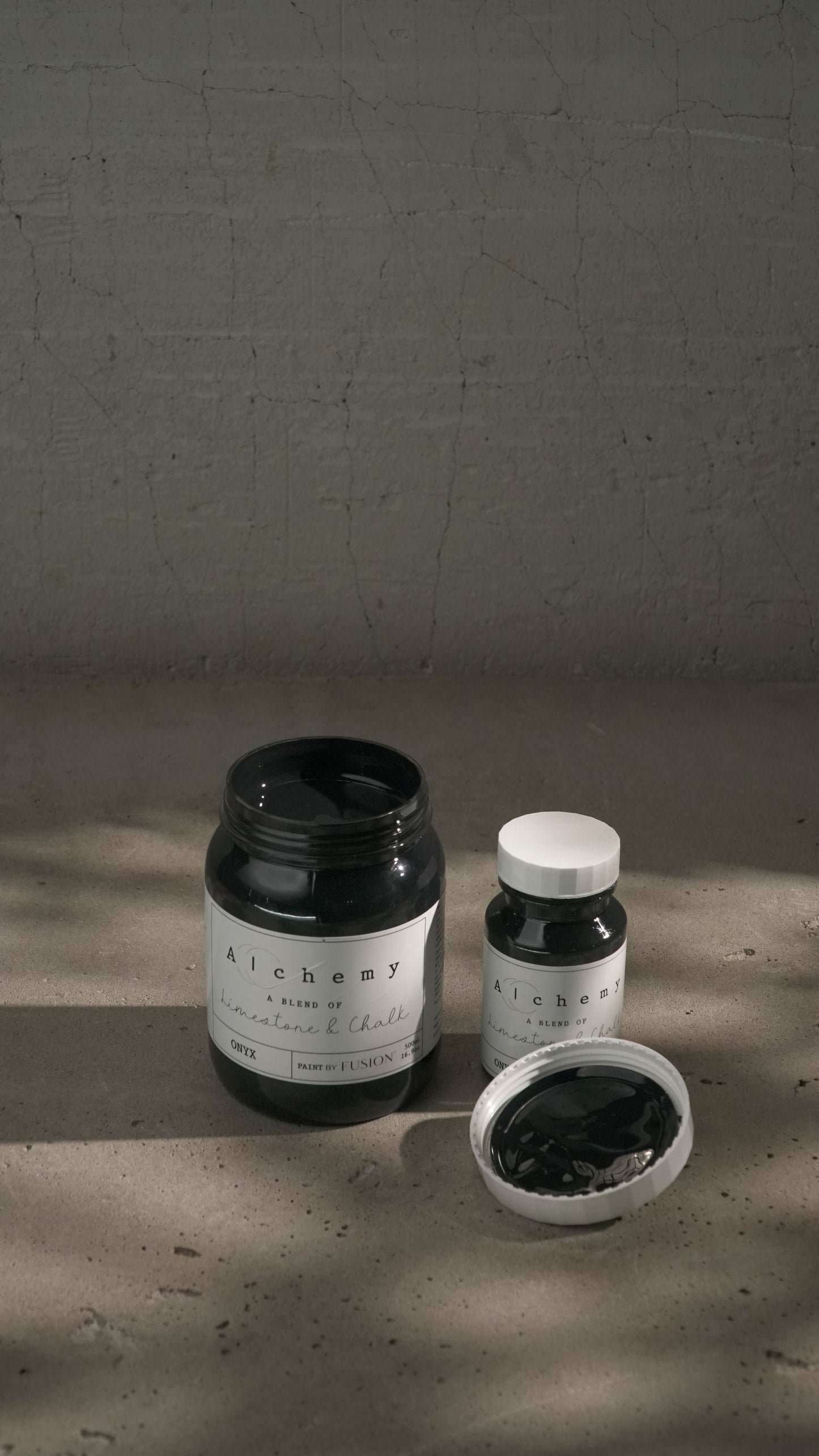 Botes Alchemy by Fusion Onyx 500ml y 120ml abiertos sobre fondo de cemento texturizado con luz lateral, pintura a la tiza negro equilibrado limestone y chalk