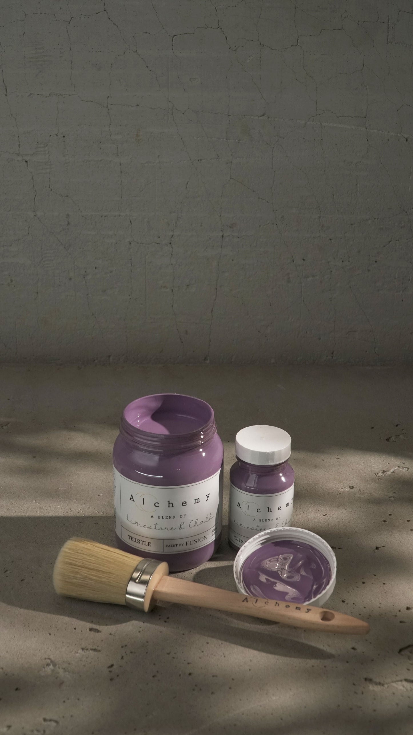 Botes Alchemy by Fusion Thistle 500ml y 120ml abiertos con brocha sobre fondo de cemento texturizado con luz lateral y sombras dramáticas, pintura a la tiza malva limestone y chalk