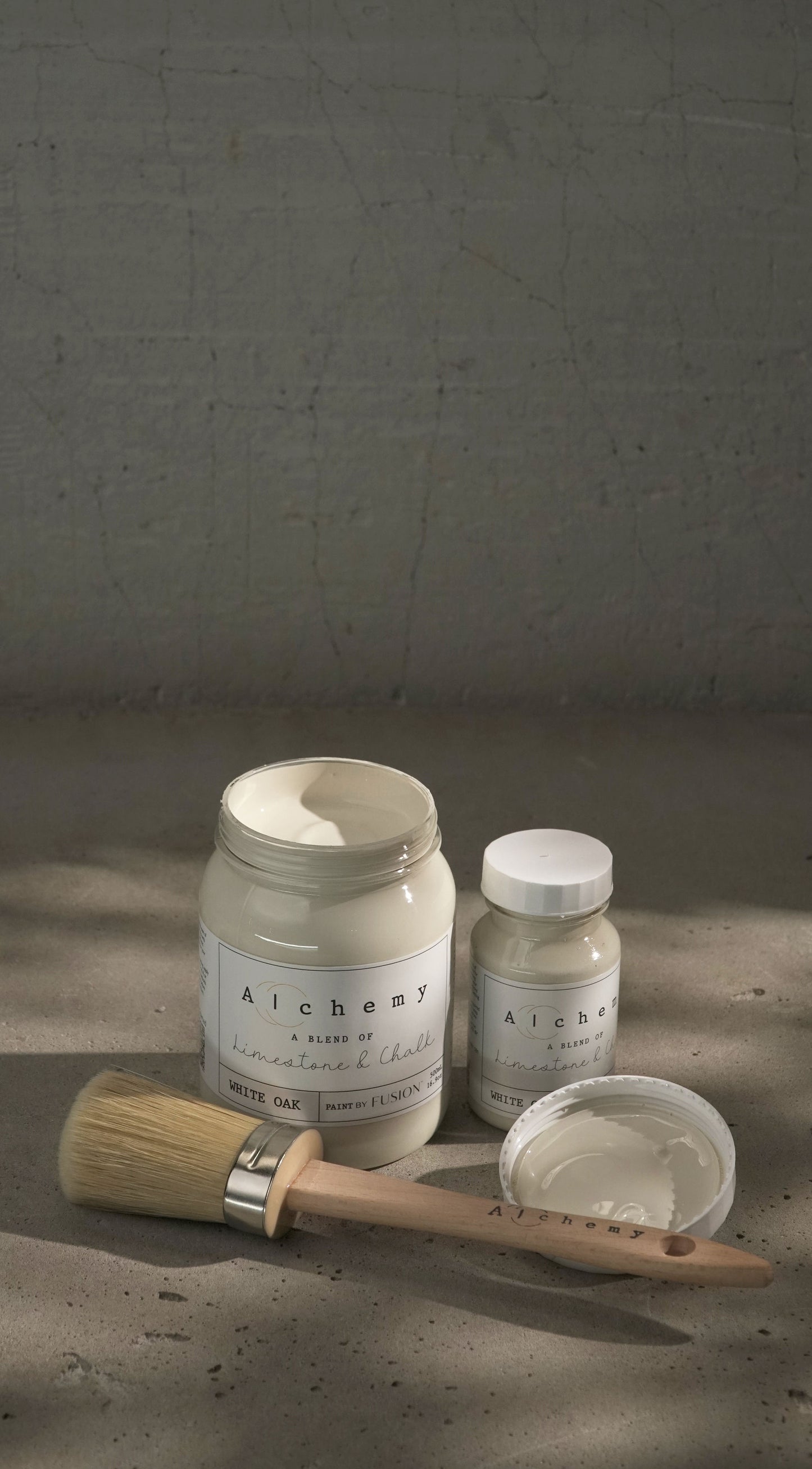 Botes Alchemy by Fusion White Oak 500ml y 120ml abiertos con brocha redonda sobre fondo de cemento texturizado con luz lateral dramática, pintura a la tiza blanco roto limestone y chalk