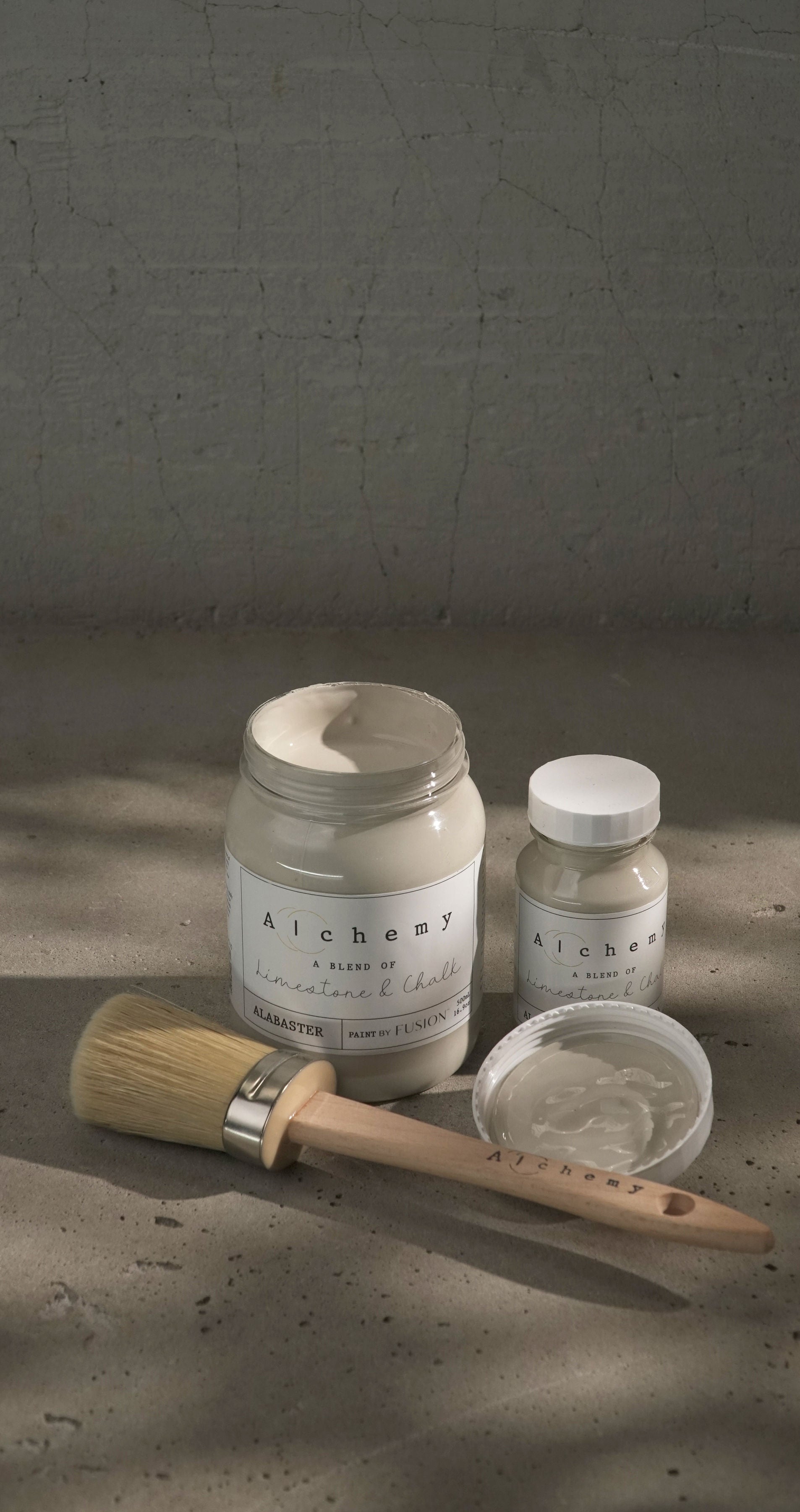 Botes Alchemy by Fusion Alabaster 500ml y 120ml con brocha sobre superficie de cemento, vista vertical, pintura a la tiza greige elegante