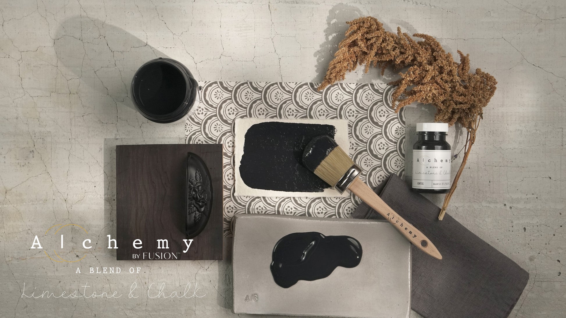 Flatlay inspiracional Alchemy by Fusion Onyx con muestra de color negro, bote 120ml, brocha redonda, tirador tallado y musgo seco sobre fondo de azulejo craquelado


