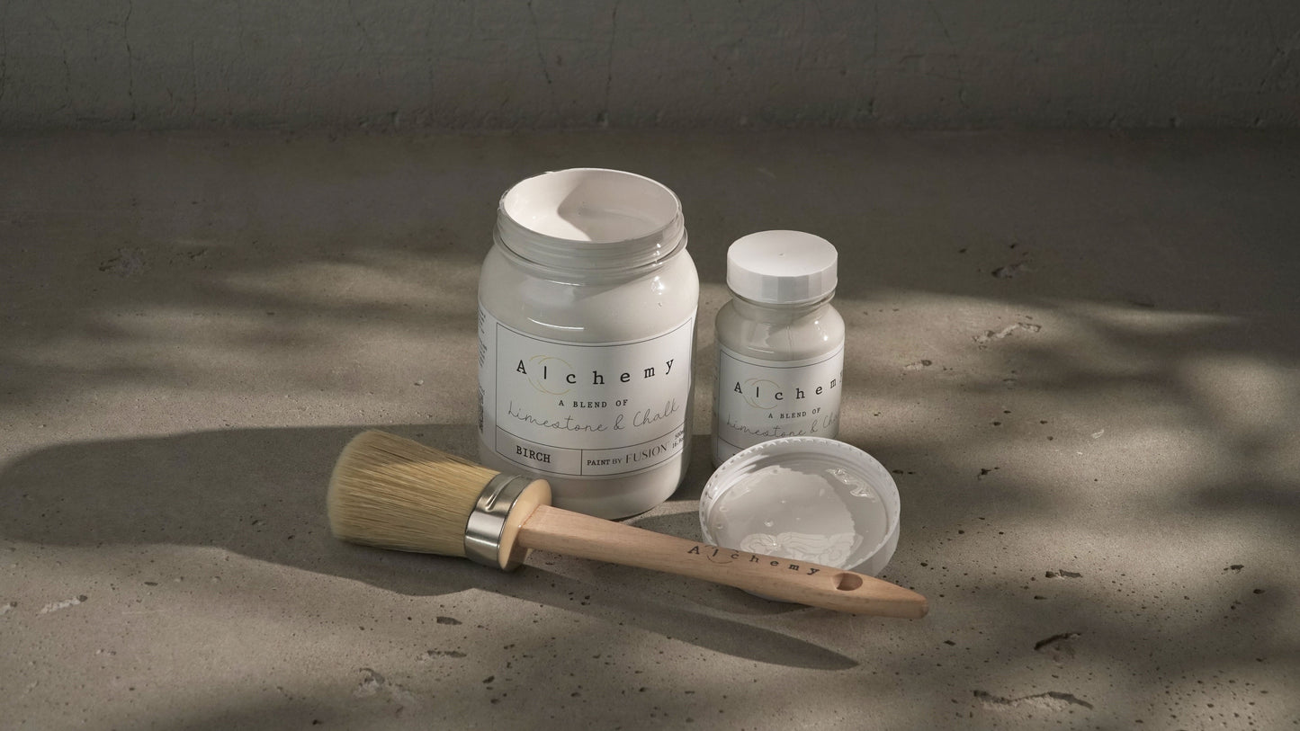 Botes Alchemy by Fusion Birch 500ml y 120ml con brocha redonda sobre superficie de cemento con luz natural, pintura a la tiza blanco roto terroso