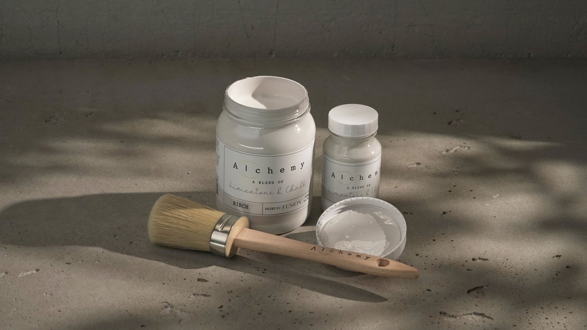 Botes Alchemy by Fusion Birch 500ml y 120ml con brocha redonda sobre superficie de cemento con luz natural, pintura a la tiza blanco roto terroso
