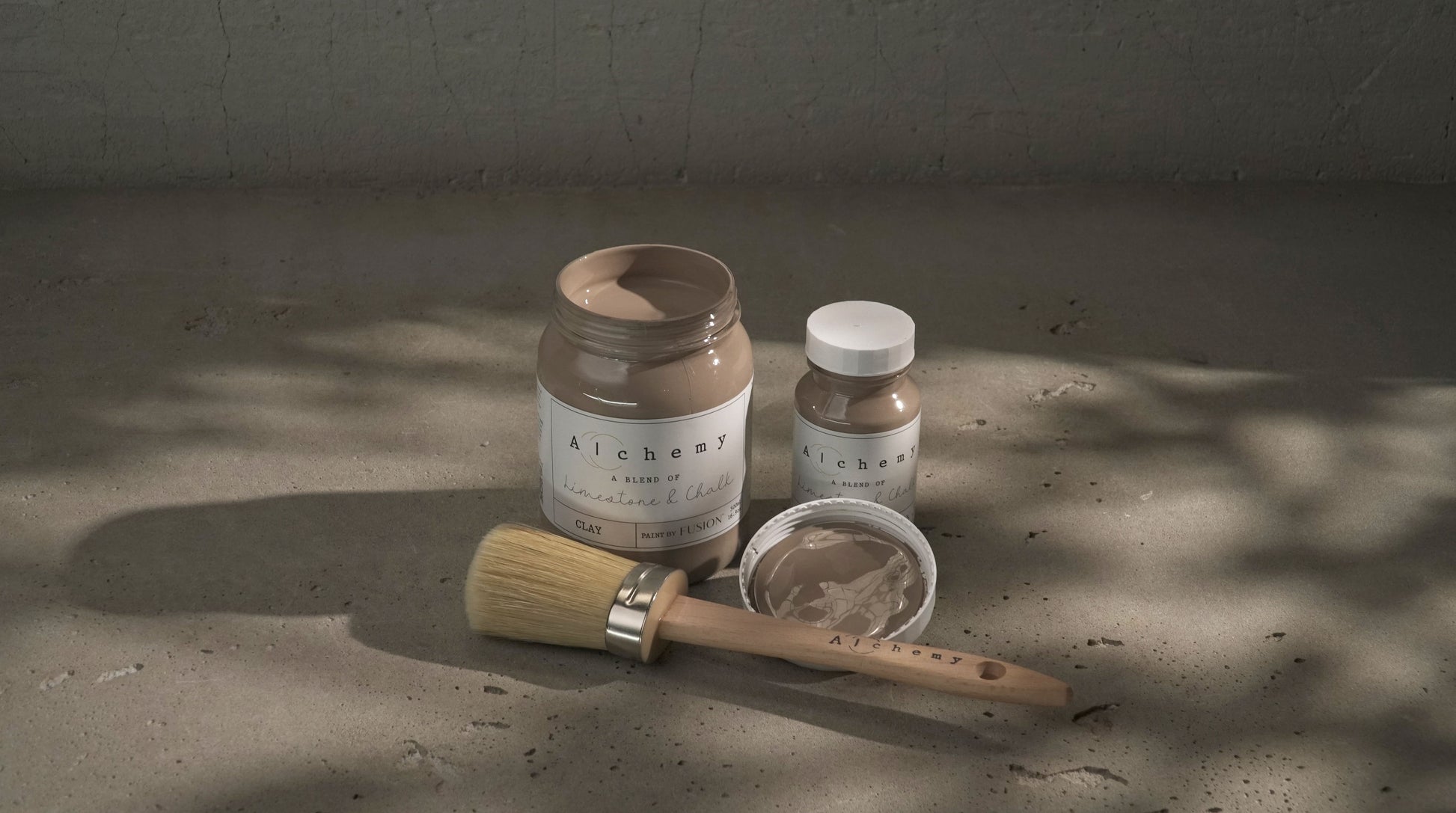 Botes Alchemy by Fusion Clay 500ml y 120ml con brocha redonda sobre superficie de cemento con sombras naturales, pintura a la tiza taupe gris cálido