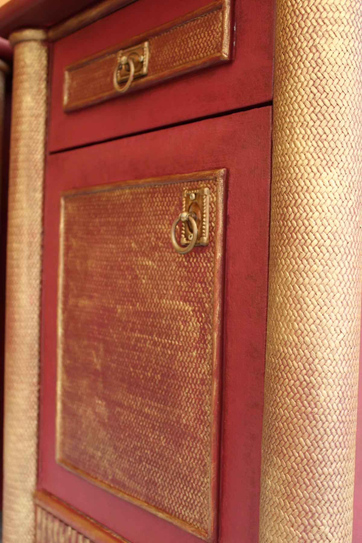Detalle de la puerta y el cajón de una mesita vintage burdeos, con paneles decorativos dorados y tiradores de anilla.