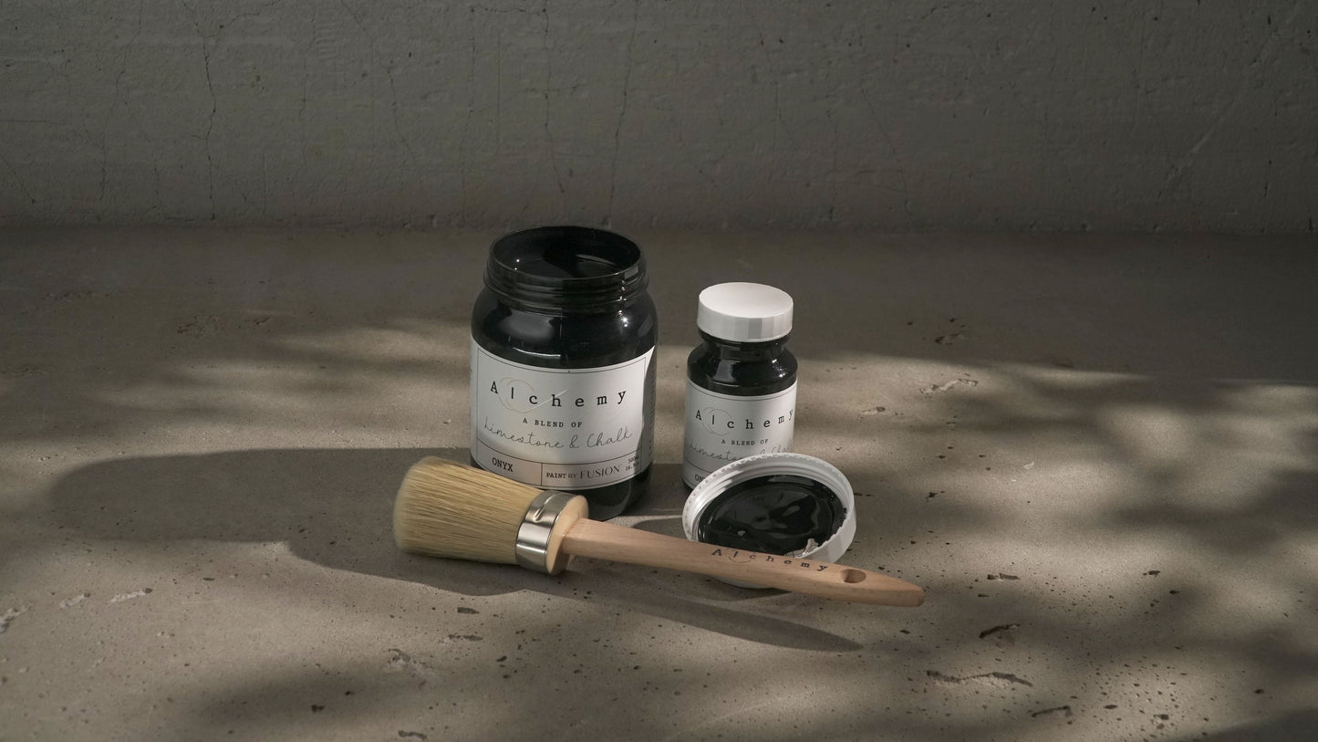 Botes Alchemy by Fusion Onyx 500ml y 120ml abiertos con brocha redonda sobre superficie de cemento con sombras de luz natural, pintura a la tiza negro puro