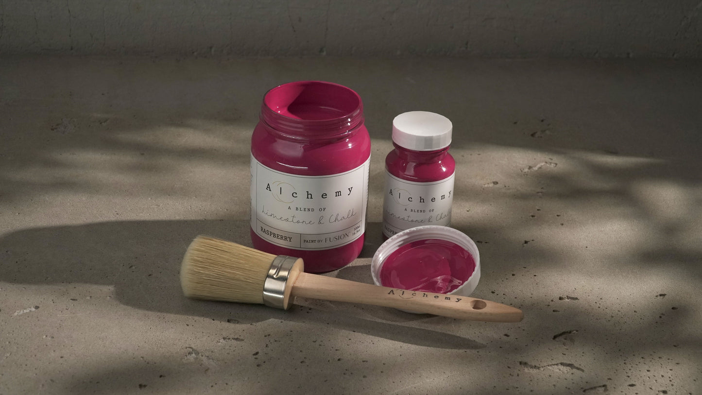 Botes Alchemy by Fusion Raspberry 500ml y 120ml abiertos con brocha redonda sobre superficie de cemento con sombras de luz natural, pintura a la tiza fucsia vibrante