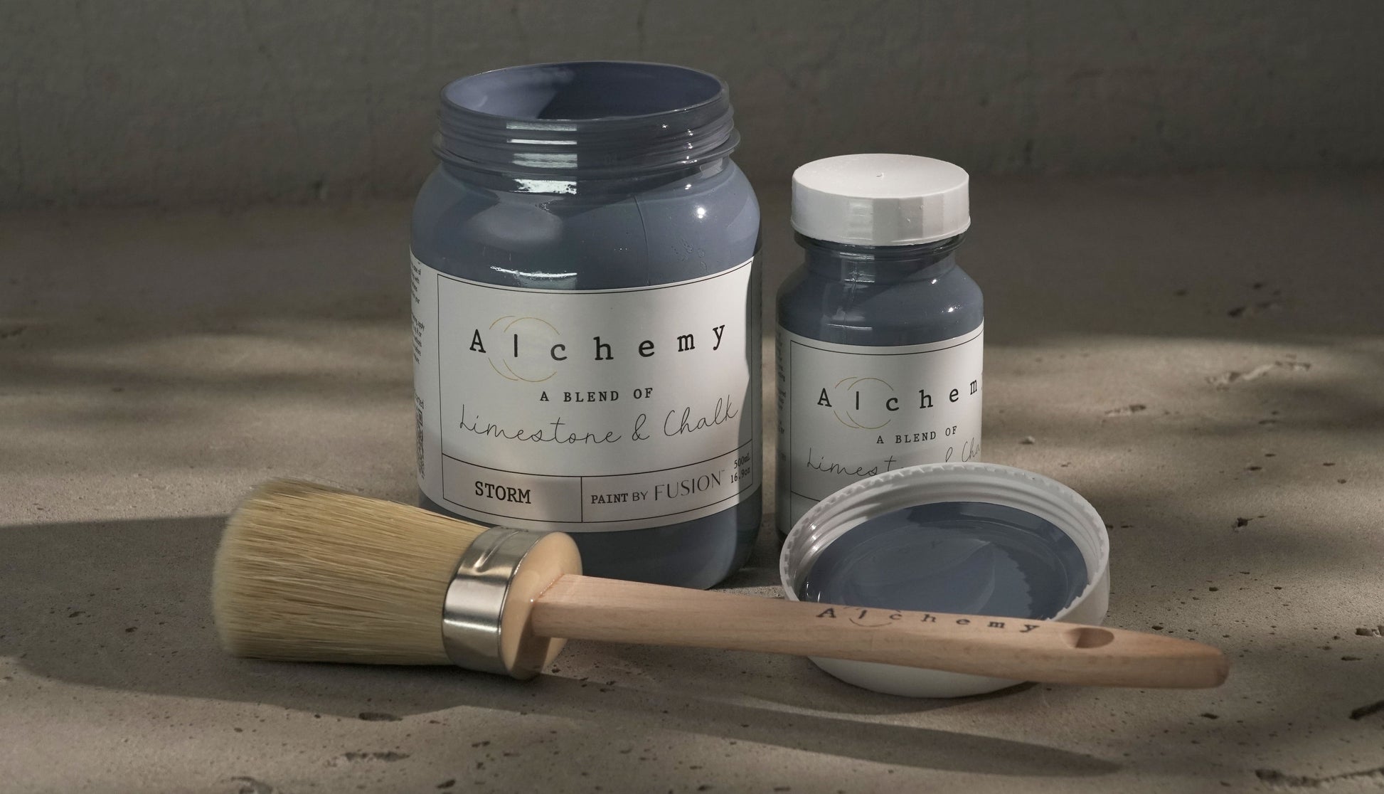 Botes Alchemy by Fusion Storm 500ml y 120ml abiertos con brocha redonda sobre superficie de cemento con sombras de luz natural, pintura a la tiza gris azulado con subtono lavanda