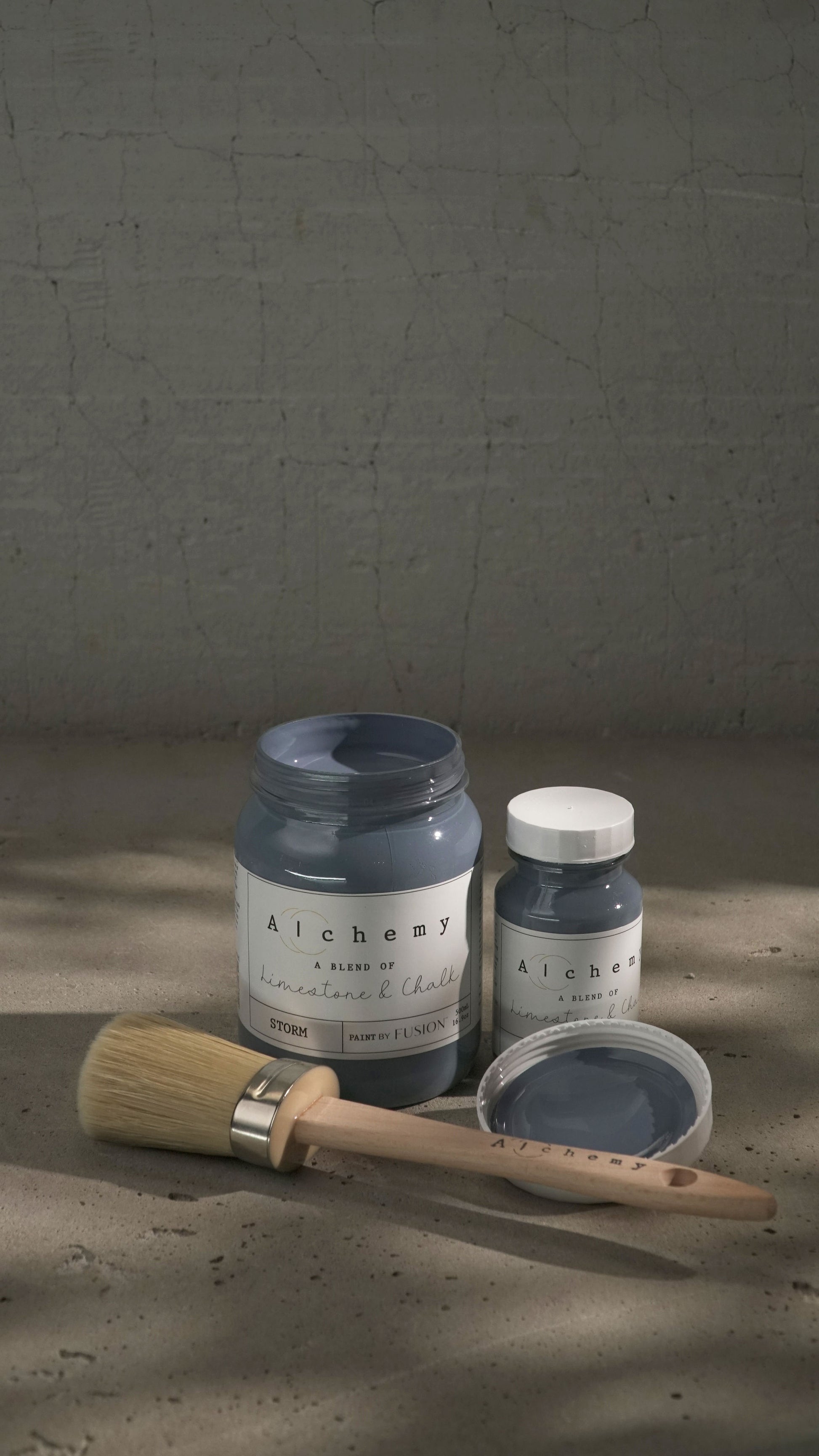 Botes Alchemy by Fusion Storm 500ml y 120ml abiertos con brocha sobre fondo de cemento texturizado con luz lateral y sombras dramáticas, pintura a la tiza gris lavanda limestone y chalk