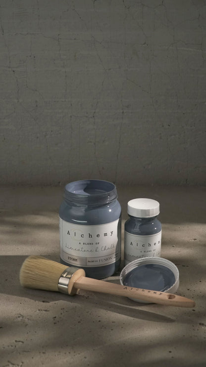 Botes Alchemy by Fusion Storm 500ml y 120ml abiertos con brocha sobre fondo de cemento texturizado con luz lateral y sombras dramáticas, pintura a la tiza gris lavanda limestone y chalk