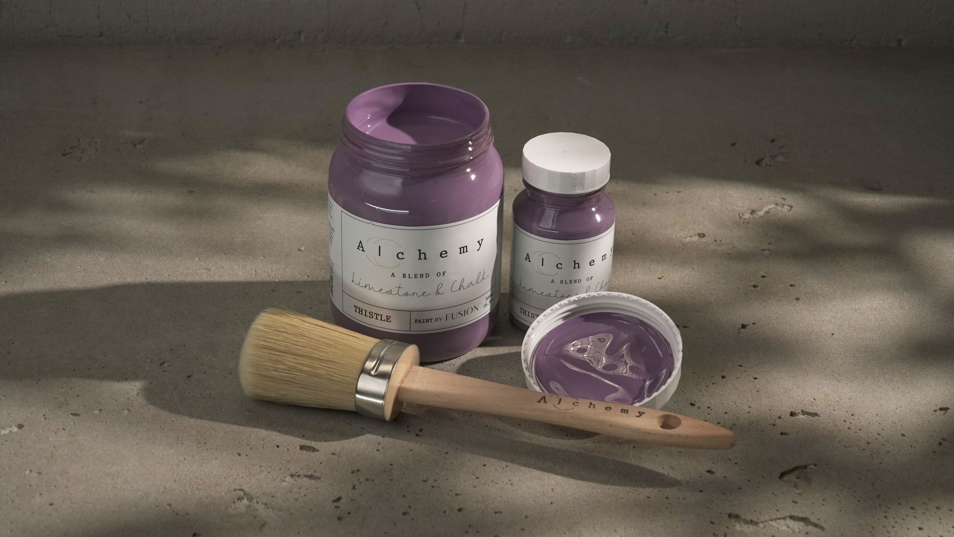 Botes Alchemy by Fusion Thistle 500ml y 120ml abiertos con brocha redonda sobre superficie de cemento con sombras de luz natural, pintura a la tiza malva profundo