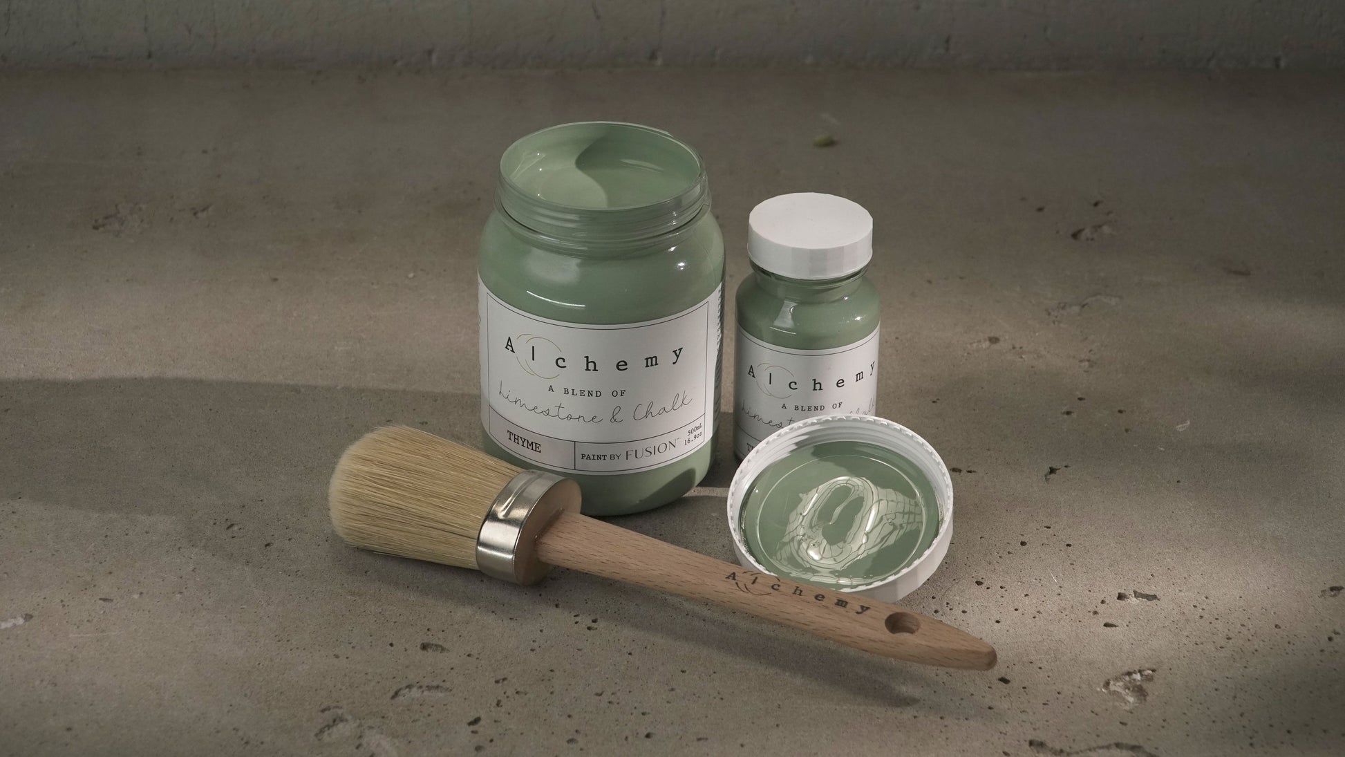 Botes Alchemy by Fusion Thyme 500ml y 120ml abiertos con brocha redonda y tapa sobre superficie de cemento con luz lateral, pintura a la tiza verde azulado fresco