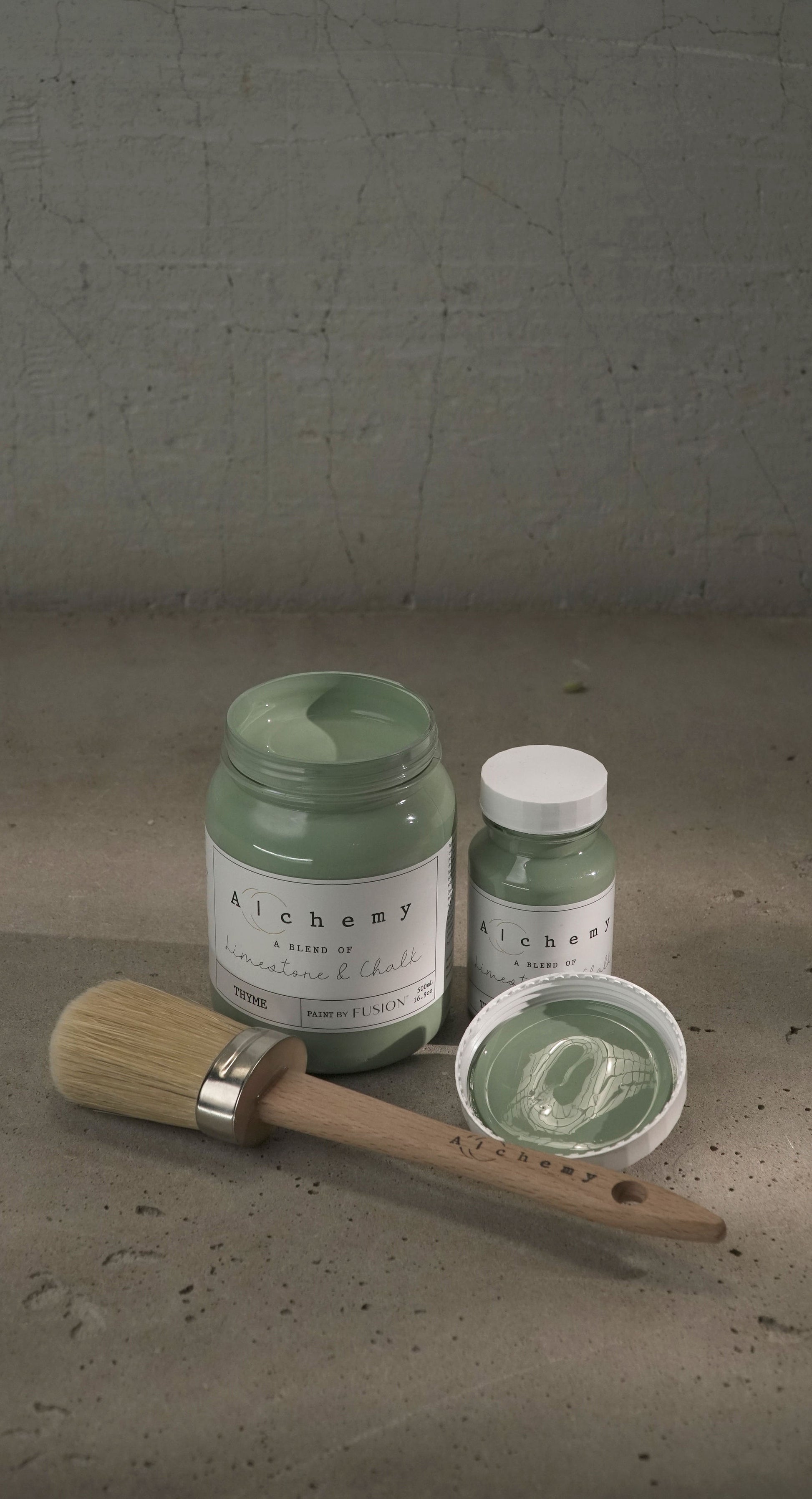 Botes Alchemy by Fusion Thyme 500ml y 120ml abiertos con brocha sobre fondo de cemento texturizado con luz cenital y sombras suaves, pintura a la tiza verde azulado limestone y chalk