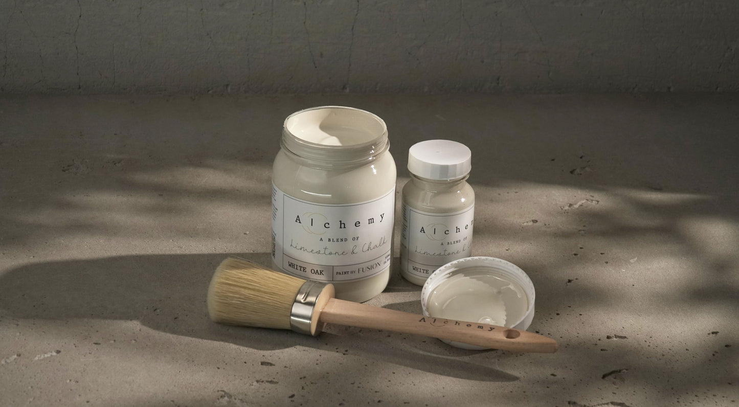 Botes Alchemy by Fusion White Oak 500ml y 120ml abiertos con brocha redonda y tapa sobre superficie de cemento con sombras de luz natural, pintura a la tiza blanco roto antiguo
