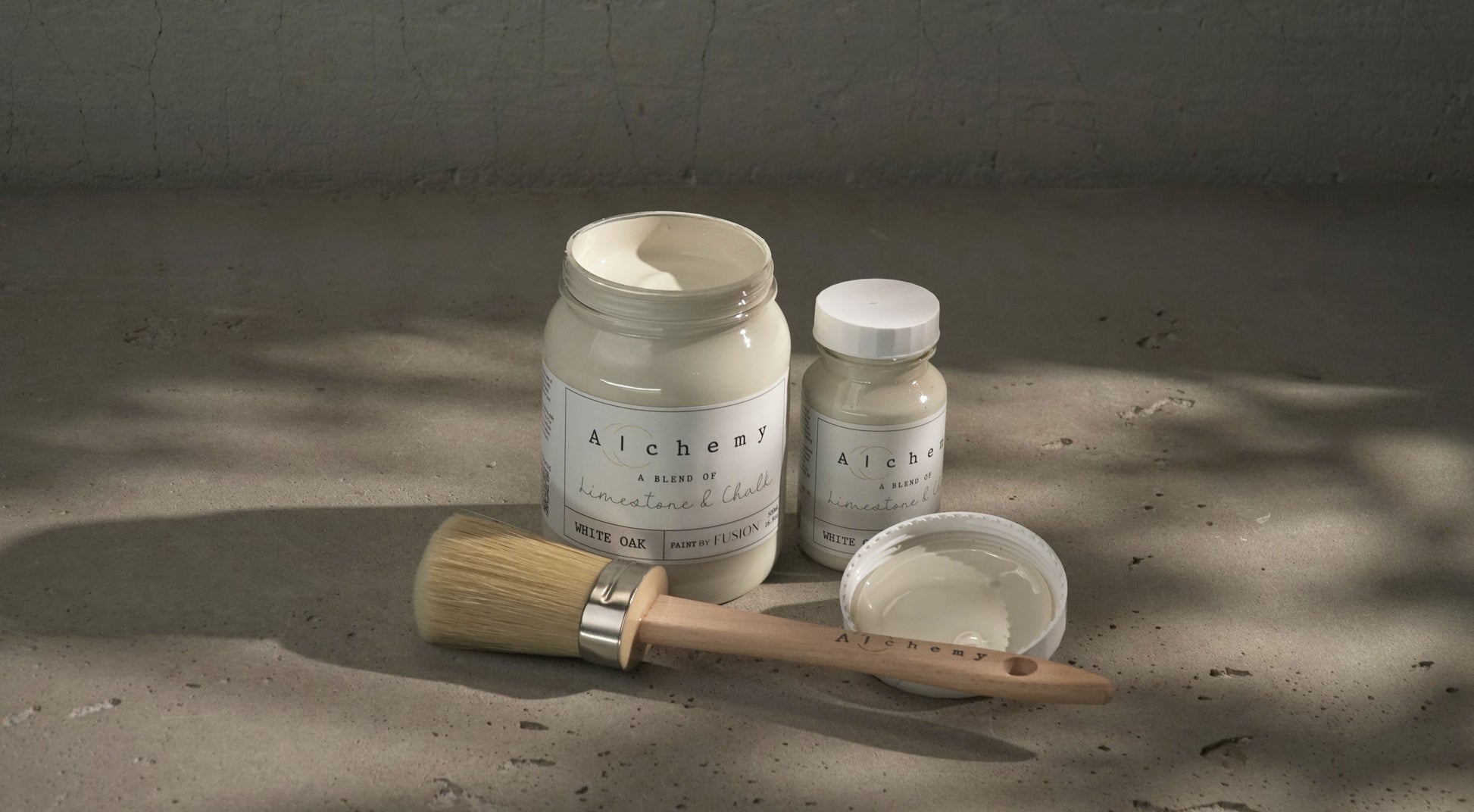 Botes Alchemy by Fusion White Oak 500ml y 120ml abiertos con brocha redonda y tapa sobre superficie de cemento con sombras de luz natural, pintura a la tiza blanco roto antiguo