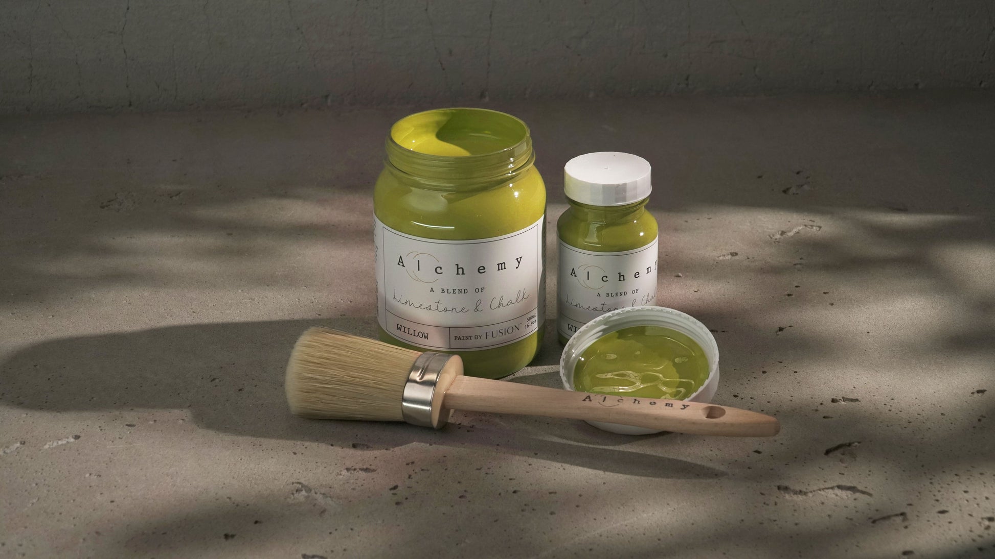 Botes Alchemy by Fusion Willow 500ml y 120ml abiertos con brocha redonda y tapa sobre superficie de cemento con sombras de luz filtrada, pintura a la tiza mostaza y chartreuse