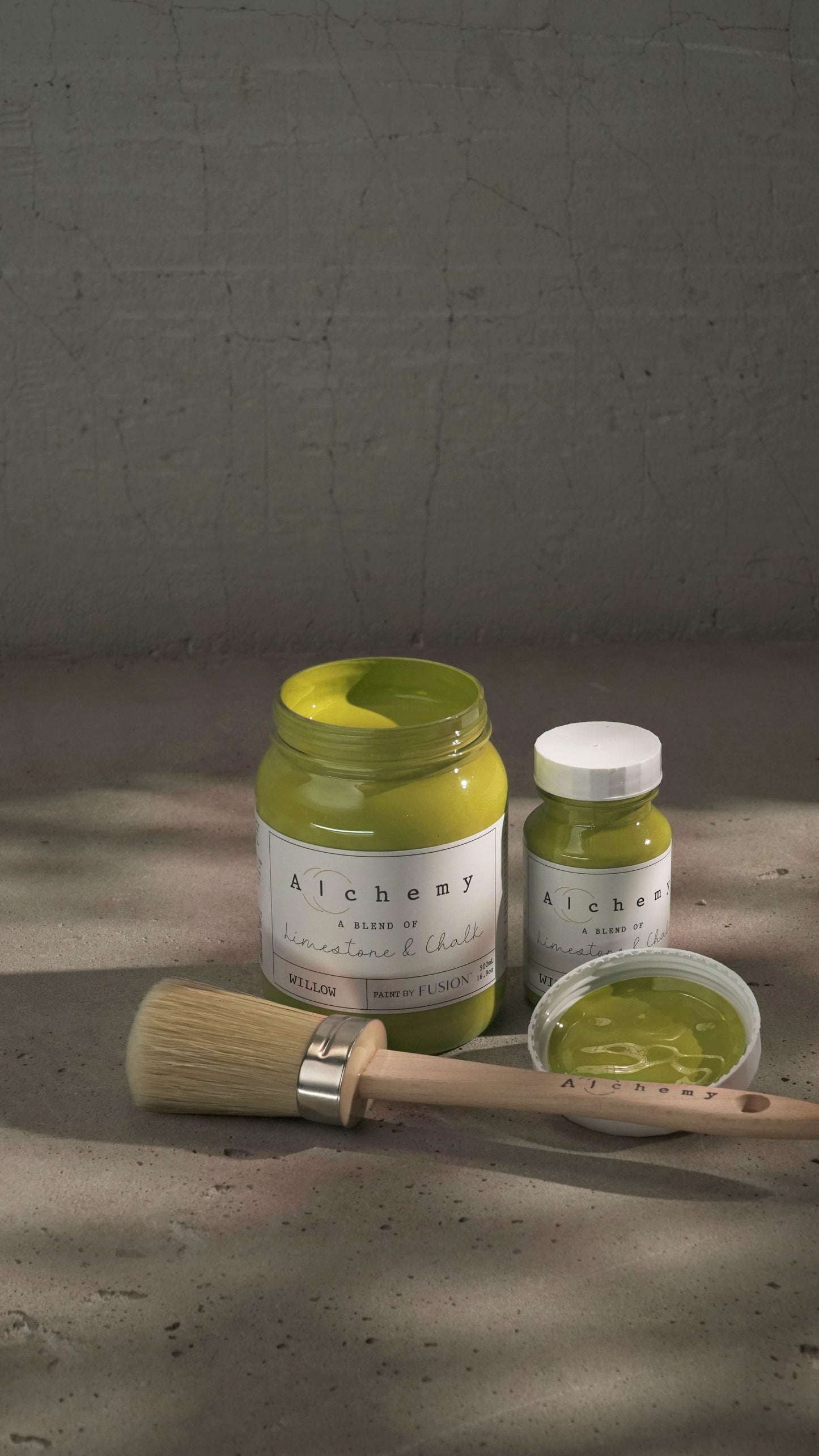 Botes Alchemy by Fusion Willow 500ml y 120ml abiertos con brocha redonda sobre fondo de cemento texturizado con luz lateral dramática, pintura a la tiza mostaza chartreuse limestone y chalk