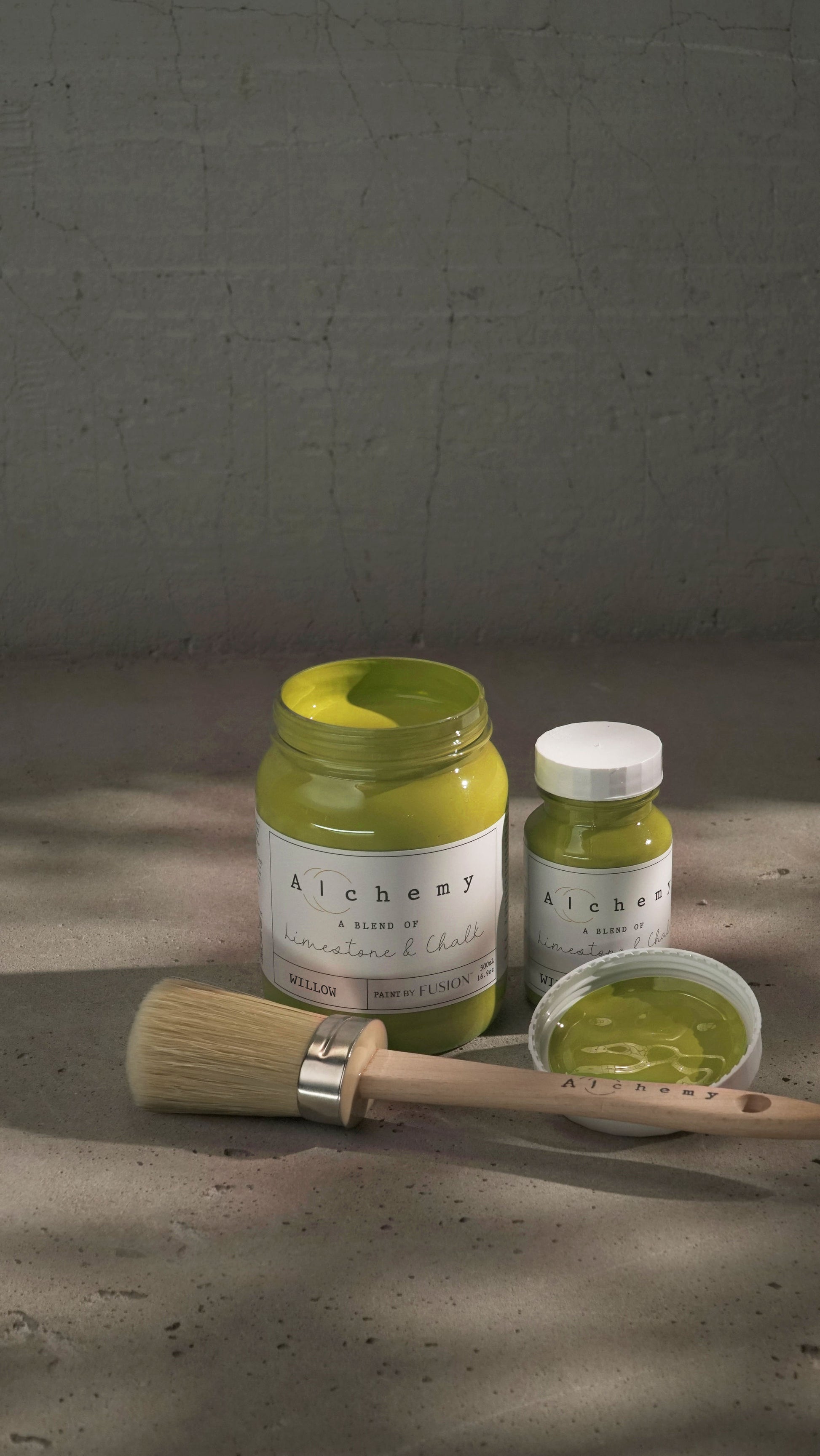 Botes Alchemy by Fusion Willow 500ml y 120ml abiertos con brocha redonda sobre fondo de cemento texturizado con luz lateral dramática, pintura a la tiza mostaza chartreuse limestone y chalk