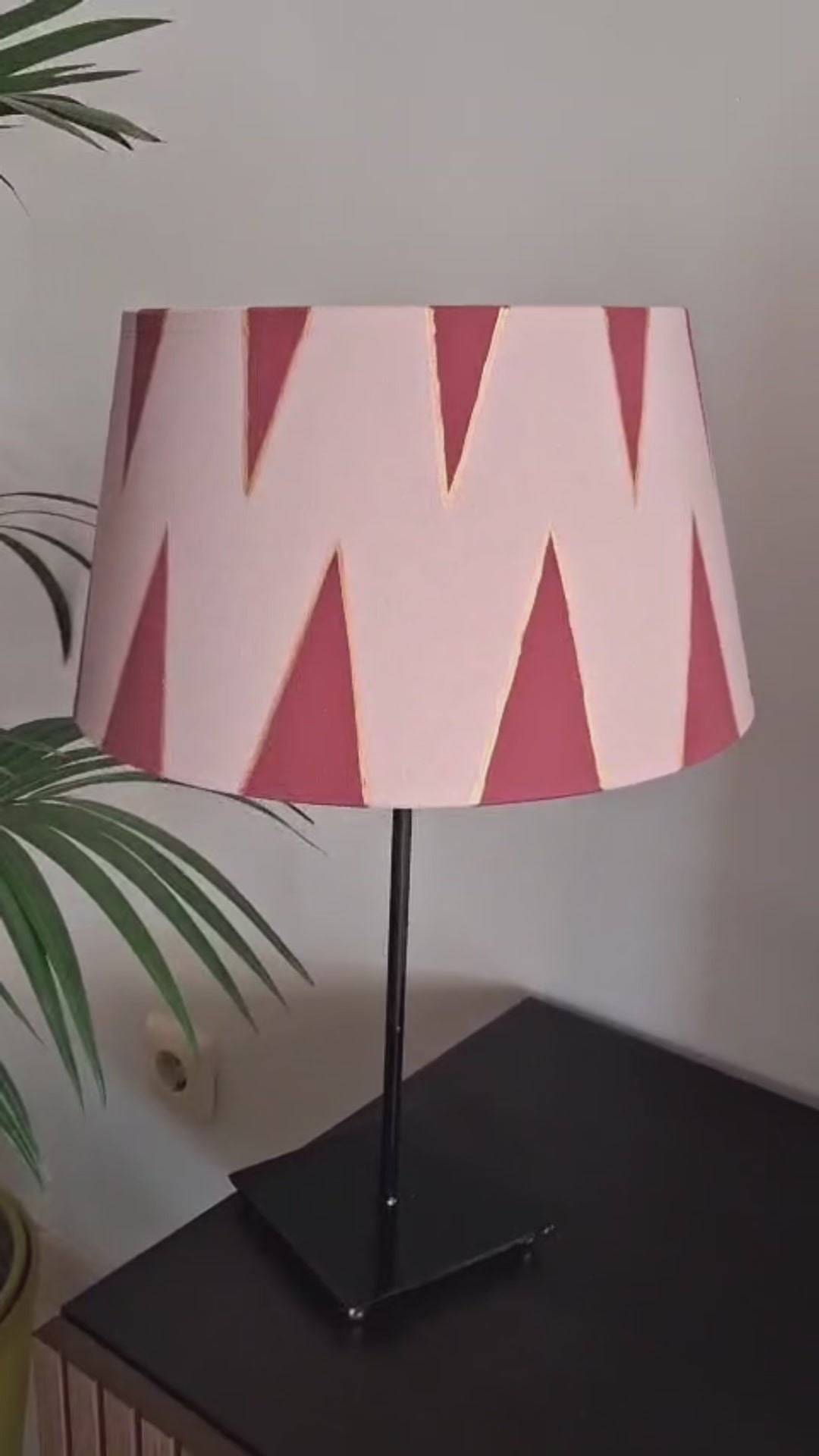 Pantalla de lámpara rosa empolvado con triángulos burdeos perfilados en dorado, presentada sobre una base negra junto a una planta.