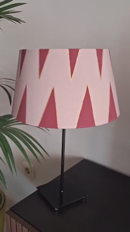 Pantalla de lámpara rosa empolvado con triángulos burdeos perfilados en dorado, presentada sobre una base negra junto a una planta.